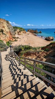 A wooden path, wooden staircase leads to a sandy beach with rocks and visitors, the still sea under