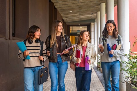 Four young women students are walking together along an outdoor university corridor, carrying books