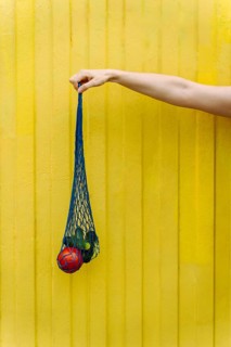 Hand holding mesh grocery bag with fresh tomatoes and cucumbers on yellow, A hand holds a blue mesh