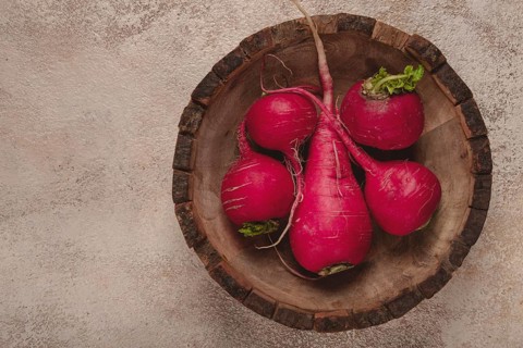 Vibrant red radishes with green tops arranged in a rustic wooden bowl on a textured surface create