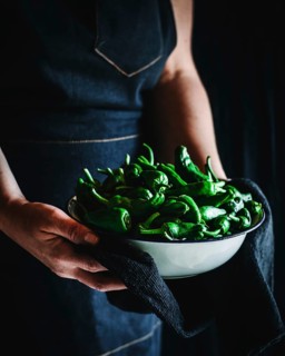 A person holds a rustic white bowl filled with fresh green peppers, wrapped in a dark cloth. The