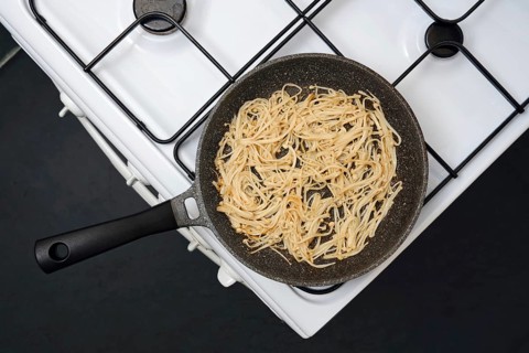 Overhead view of a frying pan with golden-brown enoki mushrooms on a gas stove, showing a homemade