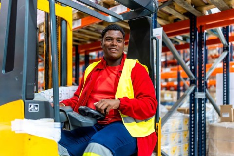 Happy young latin man driving forklift in a distribution warehouse
