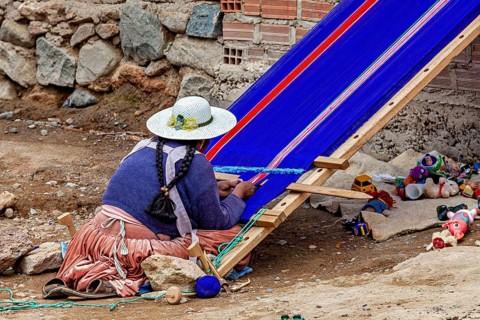 A woman in traditional garb carefully weaves on a blue loom in front of a brick wall, a woman