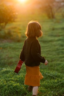 A young girl walks through a green meadow with orange trees during sunset, holding a small red