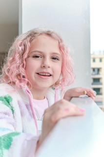 A cheerful child with pink curly hair and a colorful jacket smiles while enjoying a sunny day