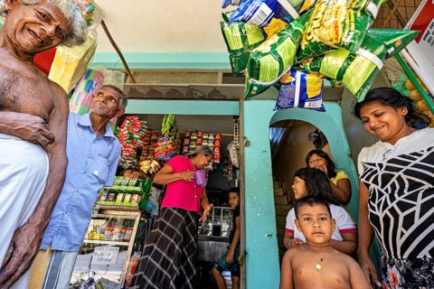People and children laugh together in front of a small shop filled with goods, a family in