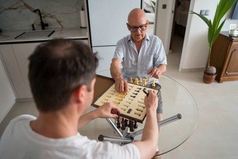 Back of unrecognizable adult son and elderly father enjoying a chess game at home, showcasing