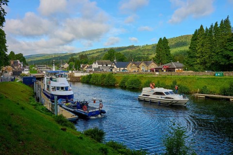 Boats sail on a river with green hills and houses in the background under a clear sky, Caledonian