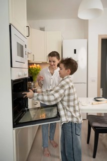 A mother and her son enjoy baking together in a bright kitchen. The son places a tray in the oven