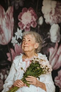 An elderly woman in a studio holds a bouquet of flowers, looking serene. She stands against a