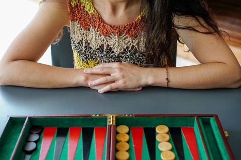 Woman sitting at a table, engaged with a backgammon board She wears a multicolored crochet top The