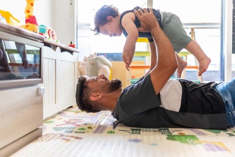 Father lying on a playmat, joyfully lifting his toddler son into the air, creating a strong bond