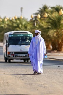 A man in traditional dress walks on a road through a desert landscape with palm trees and a bus,