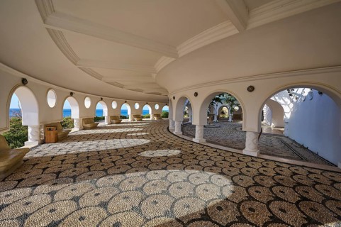 Interior of a circular building with open arches and a view of the sea, the floor is artfully
