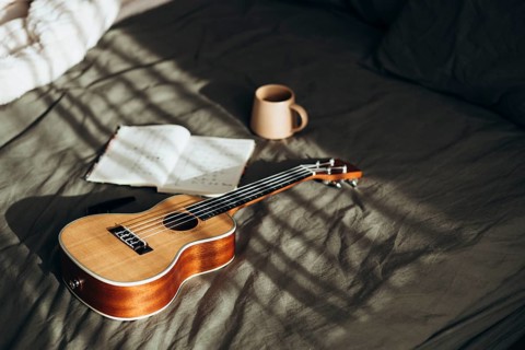 A ukulele rests beside an open notebook and a coffee mug on a softly sunlit bed This cozy scene
