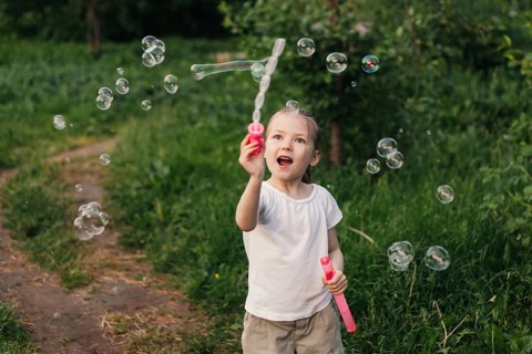 A joyful little girl plays with soap bubbles in a lush green outdoor setting. Her expression of