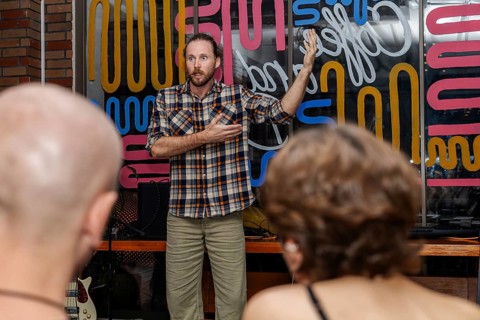 A man with a beard and plaid shirt speaks to an audience in a colorful venue. Two people with short