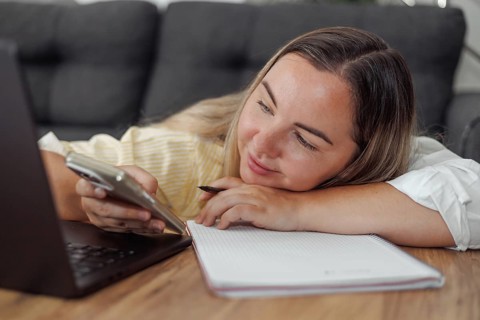 Young Caucasian woman with brown hair lying on a couch using a smartphone with a laptop and