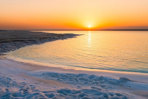 Dead Sea Jordan sunset with orange glow, calm water reflection, palm silhouettes, peaceful travel