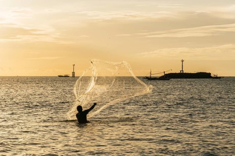 A silhouette of a fisherman casting a net into the ocean during a golden sunset. Boats are visible