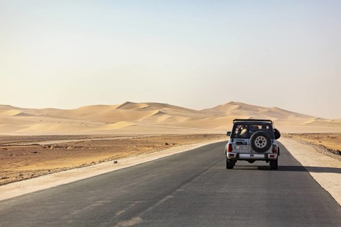 Vehicle travelling on a straight asphalt road amidst vast sand dunes under a blue sky, Driving