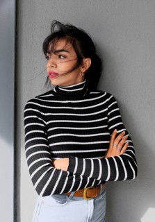 A woman in a black and white striped turtleneck sweater leans against a gray wall. She crosses her