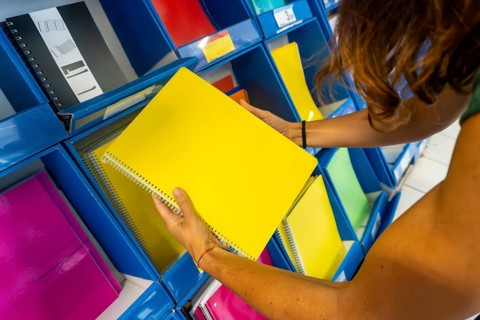 A woman is holding a yellow spiral notebook in a store. The notebook is on a shelf among other