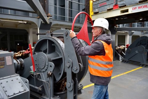 A worker wearing helmet and vest operates a large machine on a ship in an industrial environment,