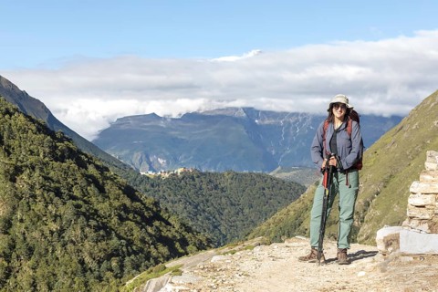 A woman smiles while hiking the Everest trail, surrounded by lush green mountains and clear blue