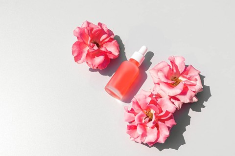 Top view of a rose-tinted serum bottle surrounded by vibrant pink roses, set against a smooth white