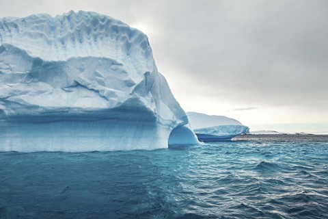 A large iceberg rises majestically out of the blue water under a grey sky, icebergs in the Southern