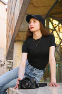 A woman in casual attire, with long hair and a cap, leans against a city building holding a camera.