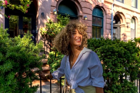 Young woman with curly hair smiling in bright sunlight, experiencing urban summer life on a new