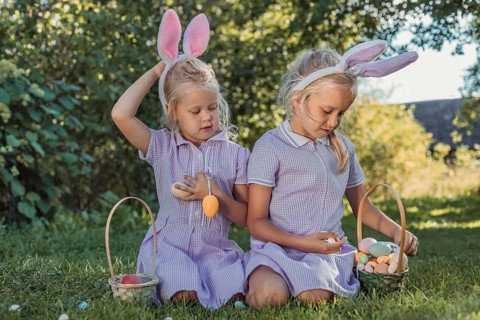 Two young girls, wearing bunny ears and dresses, search for colorful Easter eggs in a sunny garden