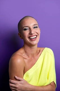 A middle-aged woman with a shaved head beams with a bright, genuine smile. She wears a vibrant
