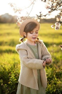 A young girl, wearing a beige hat and sweater, admires almond blossoms on a sunny spring day