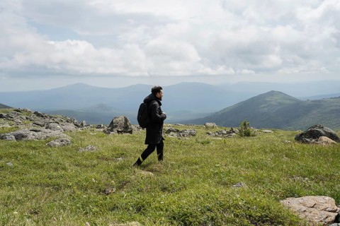 A lone hiker walks across a grassy mountain terrain, surrounded by rocky outcrops and distant peaks