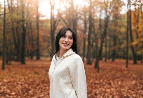 A woman wearing a cozy sweater stands in a forest filled with vibrant autumn foliage. The ground is