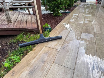 Wet wooden deck with a squeegee and green grass. A wooden patio is visible in the background,