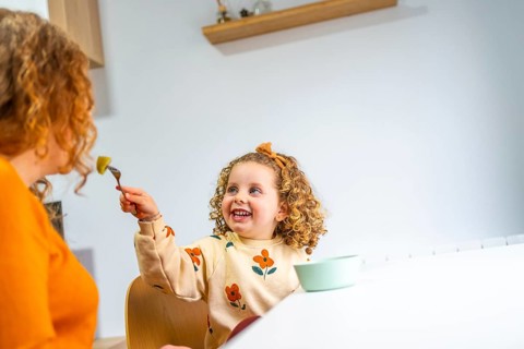 Playful and smiling caucasian school girl eating at home next to her mother