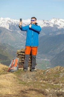 A man, wearing a blue jacket and orange pants, takes a selfie while trekking on a picturesque trail
