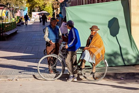 Three people stand on a street in sunny weather, one on a bicycle in an urban environment, cyclists