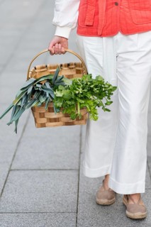 Fashion woman in white pants and red jacket holding a wicker basket filled with fresh herbs,