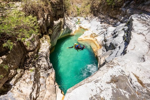 A canyoneer is captured in mid-flight in a dynamic jump into a shimmering turquoise pool surrounded