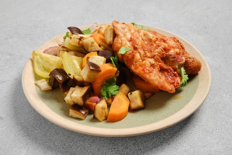 Grilled chicken piece sits on plate next to mix of roasted vegetables including carrots, eggplant