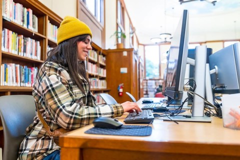 Young millennial woman wearing a beanie and plaid shirt sitting at a computer workstation, actively