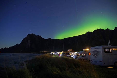 Motorhomes park on the beach under a clear night sky with northern lights and dark mountains,