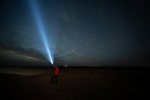 A man with flashlight in a star filled night sky over a Merzouga desert landscape. The faint glow