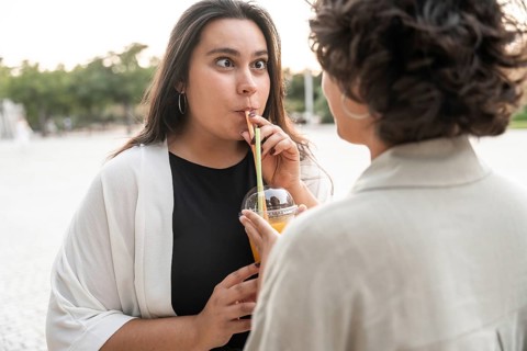 A lesbian couple enjoys a playful moment while sharing a refreshing drink outdoors. The pair exudes